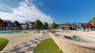 Located at 1920 Exchange Dr in Greenville, North Carolina, this outdoor amenity complex delivers a resort-caliber experience designed for active recreation and relaxed leisure. At the heart of the space is a generously sized, freeform swimming pool with clear turquoise water, complemented by a distinctive checkered-tile deck that adds a bold visual character to the surroundings. A central raised hot tub serves as a focal point within the pool area, while a separate shallow pool with a volleyball net provides additional recreational options.

Modern sculptural lounge chairs are arranged throughout the pool deck, offering ample space for sunbathing and relaxation. Covered pergola structures provide shaded retreats, and mature palm and deciduous trees add natural beauty and seasonal color to the landscape. Lush artificial and natural turf areas are thoughtfully integrated throughout the grounds, creating green gathering spaces between amenity zones.

An outdoor fitness area features a multi-station obstacle and training rig, catering to residents seeking active workouts in the open air. Tucked beneath a mature shade tree, a hanging wicker egg chair offers a tranquil nook for quiet relaxation. The well-maintained grounds are enclosed by decorative iron fencing, with multi-story residential buildings framing the perimeter and contributing to the community's cohesive aesthetic.