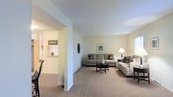 This single-floor residence offers a thoughtfully designed layout that balances comfort and functionality. The living room serves as the heart of the home, featuring soft, neutral wall tones and plush wall-to-wall carpeting that creates a warm and inviting atmosphere. The space is generously proportioned, comfortably accommodating a full seating arrangement centered around a sleek coffee table — ideal for both relaxing and entertaining. A decorative wall accent adds a subtle artistic touch to the room's understated aesthetic. An open archway connects the living area to an adjacent kitchen or dining space, enhancing the sense of openness and flow throughout the floor plan. The kitchen area, visible through the passthrough, features a breakfast bar or counter ledge that further encourages an open, connected feel between rooms. Natural light filters through the space, complementing the bright, clean ceiling and light-colored walls. The overall design reflects a practical yet comfortable living environment, with a cohesive color palette of soft beiges and grays that lend the home a calm, neutral character well-suited to a variety of interior styles and furnishings.
