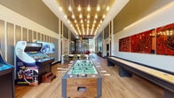 This expansive amenity lounge is designed to impress, offering a thoughtfully curated environment for recreation and socializing. The space is defined by soaring double-height ceilings and warm hardwood-style flooring that runs throughout, creating an open and energetic atmosphere. A striking cascade of Edison-style pendant lights illuminates the central game area, which features a full-size foosball table, a shuffleboard table, and a classic arcade machine — all set against a backdrop of bold, large-scale wall art, including a vibrant red circuit-board-inspired triptych and expressive abstract paintings. A dedicated lounge area offers a generous sectional sofa accented with colorful throw pillows, an artistic gold sculptural coffee table, and a dramatic woven pendant light fixture, all framed by curated artwork. Floor-to-ceiling windows at the far end of the space flood the interior with natural light and provide views of lush greenery outdoors. A sleek wet bar with rich dark wood cabinetry, white countertops, a stainless steel under-counter refrigerator, and a wine cooler adds a functional entertaining element. Adjacent to the lounge, a fitness center is accessible through a glass-door entry, flanked by eye-catching yellow sculptural chairs. The overall design blends contemporary style with a vibrant, social character.