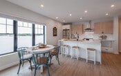 This model unit at 10 Congress St is a well-appointed 2-bedroom, 2-bathroom residence that blends contemporary design with a coastal aesthetic across a single floor. The open-concept kitchen is a standout feature, showcasing quartz countertops, a large island, two-tone cabinetry with light wood upper cabinets and crisp white lower cabinets, a white subway tile backsplash, stainless steel appliances including a range with a stainless hood, and matte black hardware throughout. Light hardwood flooring runs seamlessly through the entire unit. The living area is bright and welcoming, with a large sliding glass door opening to a private balcony that offers water views, and a wall-mounted mini-split system for climate control. The primary bedroom is generously sized, featuring recessed lighting, a walk-in closet, natural light from a window with water views, and its own en-suite bathroom with a glass-panel shower and modern fixtures. The second bedroom also enjoys water views through large black-framed windows. A second full bathroom provides a soaking tub and shower combo, a floating vanity, and matte black accents. The unit's coastal-inspired decor and neutral palette create a serene, refined atmosphere throughout.