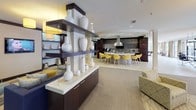 This single-floor amenity space is designed with a contemporary, hospitality-inspired aesthetic that prioritizes both comfort and social interaction. The open-plan layout flows seamlessly between distinct functional zones, creating a cohesive and inviting environment throughout.

The main lounge area is anchored by a grouping of patterned armchairs arranged around a round white coffee table, centered beneath a large cylindrical yellow pendant light that adds a vibrant focal point to the neutral palette. Light-toned carpeting with a subtle organic motif defines this seating zone, while light tile flooring extends into the surrounding areas.

A standout architectural feature is the pair of large industrial-style garage doors that open to an outdoor terrace, where additional bar-height seating and views of lush landscaping are visible. This indoor-outdoor connection allows abundant natural light to flood the interior.

A dark-toned shelving unit displaying a curated collection of white ceramic vessels acts as a stylish room divider, separating the lounge from a full kitchen area. The kitchen features dark cabinetry, mosaic tile backsplash, stainless steel appliances, a range hood, and a counter lined with bold yellow bar stools.

Adjacent areas include a more intimate lounge with a wall-mounted television, built-in display shelving, and a gallery-style corridor showcasing large-scale artwork. The overall ambiance is polished, modern, and thoughtfully appointed.