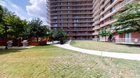 This space captures a single-floor bathroom unit located within a tall multi-story residential building. The exterior of the building reveals a classic brick-and-balcony façade rising multiple stories, complemented by well-maintained green lawns, mature trees, concrete walkways, and an on-site playground area — offering a sense of community and outdoor accessibility.

The bathroom itself is finished in soft, warm neutral tones throughout, with cream and beige walls creating a calm, understated atmosphere. The vanity features a wood-grain cabinet base with a white integrated sink basin, chrome dual-handle faucet hardware, and a framed rectangular mirror mounted above with a wood-toned border that adds a touch of warmth. A GFCI-protected electrical outlet with a brass-finish cover plate is conveniently positioned near the vanity, consistent with the brass and gold-toned hardware accents found throughout the space, including the light switch plate.

The toilet area is tucked neatly into its own alcove, with a small decorative wall-mounted shelf and framed artwork displayed above, adding a personal touch to the functional space. A bar-style towel holder is also present. The overall layout is efficient and well-organized, making practical use of the available space while maintaining a cohesive, neutral aesthetic throughout.