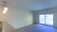 This single-floor space offers a clean, open layout designed to maximize comfort and functionality. The main living area is finished with wall-to-wall carpeting and features smooth, neutral-toned walls that create a bright and welcoming ambiance. A vaulted ceiling adds a sense of openness and volume to the room, enhancing the overall sense of space.

A standout feature of the living area is the large sliding glass door, which floods the interior with an abundance of natural light and provides direct access to an outdoor balcony. The balcony overlooks a lush, green exterior, offering pleasant views of surrounding trees and neighboring structures. This seamless indoor-outdoor connection enhances both the livability and the visual appeal of the space.

The flooring transitions from carpet in the main living area to what appears to be a harder surface near the entryway, providing a practical delineation between zones. The neutral color palette throughout the space offers a versatile backdrop, making it easy to personalize with furnishings and décor. Overall, the space presents a straightforward, light-filled environment with practical finishes and a notable connection to the outdoors.
