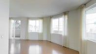 This single-floor space offers a clean, open layout designed to maximize comfort and functionality. The main living area is a standout feature, showcasing warm hardwood flooring that runs throughout, lending the space a sense of warmth and continuity. Two large windows dressed with elegant sheer cream curtains allow an abundance of natural light to filter in, creating a bright and airy atmosphere throughout the day. The windows frame views of the surrounding urban landscape, including neighboring buildings and greenery, providing a pleasant connection to the outside environment. The neutral white walls and white baseboards complement the light tones of the curtains and flooring, contributing to a clean, cohesive aesthetic. The layout of the main room is open and versatile, lending itself to a variety of furniture arrangements and uses. The ceiling is smooth and well-proportioned, adding to the sense of spaciousness without feeling overwhelming. Overall, the space presents a simple yet refined interior that balances natural light, open space, and understated design elements, making it a functional and welcoming environment suitable for a range of purposes.