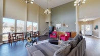 This single-floor space makes a strong first impression with its dramatically high ceilings and an open, airy layout that seamlessly connects multiple functional areas. The soaring walls are painted in a warm neutral tone, contrasted by a sage green accent wall that anchors the kitchenette and lounge zone, lending the interior a welcoming, layered character.

The main lounge area is generously proportioned, furnished with a large sectional sofa, vibrant magenta accent chairs, and a colorful area rug over rich dark hardwood flooring. An elegant chandelier overhead adds a refined touch to the space. Adjacent to the lounge, a compact kitchenette offers wood cabinetry, a double basin sink, a dishwasher, and countertop appliances, providing practical amenities within the communal setting.

A dedicated dining area near the expansive floor-to-ceiling windows features a tall bistro-style table with bar seating, offering pleasant outdoor views of surrounding greenery and neighboring structures. Additional seating, including a woven accent chair, is positioned to take full advantage of the abundant natural light that floods the room through multiple large windows.

An entryway corridor leads to exterior doors and additional rooms, with a coffee station and decorative console table contributing to the welcoming, community-oriented atmosphere of the space.