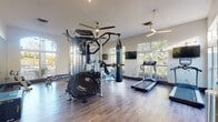 Located at 16401 Chenal Valley Dr in Little Rock, Arkansas, this fitness center is a single-floor facility designed to support a wide range of workout routines. The space features soaring vaulted ceilings that contribute to an open, airy atmosphere, while an abundance of large windows along multiple walls flood the interior with natural light and offer views of the surrounding tree-lined landscape.

The flooring throughout is a durable wood-look surface that complements the clean, neutral wall tones, creating a modern yet welcoming environment. Overhead, contemporary linear light fixtures and ceiling fans ensure the space remains well-lit and comfortable during any workout.

The facility is comprehensively outfitted with diverse equipment to accommodate both cardio and strength training. Cardio offerings include treadmills, elliptical trainers, a stationary upright bike, and a recumbent bike, each positioned near windows for an engaging workout experience. Multiple wall-mounted flat-screen TVs are positioned throughout the room for added entertainment.

For strength training, the space features a multi-station cable machine, a mini trampoline, a heavy punching bag, a full rack of dumbbells in varying weights, plyo boxes, and yoga mats. A large floor-to-ceiling mirror on one wall enhances the sense of space and allows users to monitor their form. The overall layout is thoughtfully organized, providing ample room to move freely between equipment.