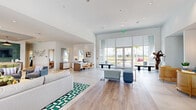 This contemporary single-floor clubhouse is thoughtfully designed to foster community and comfort through a series of well-appointed, interconnected spaces. The main gathering area is expansive and airy, featuring soaring ceilings, recessed lighting, and wide-plank wood-style flooring that transitions seamlessly into large-format tile. Floor-to-ceiling glass doors and windows flood the interior with abundant natural light while offering unobstructed views of the surrounding outdoor landscape, including swaying palm trees. Modular seating arrangements with modern ottomans and accent chairs in muted blues and grays create flexible, casual gathering zones throughout the open floor plan. A dedicated lounge area showcases a rich cognac leather banquette accented with vibrant teal throw pillows, a marble-top accent table, and a woven rattan armchair, all set against a sophisticated panel-molded accent wall adorned with contemporary artwork and wall sconces. Built-in shelving with a mounted flat-screen television provides a media-centric focal point. The kitchen and bar area is a standout feature, boasting a dramatic full-wall backsplash in glazed emerald green tile, a white quartz countertop island, wood-toned cabinetry, a built-in wine cooler, and pendant lighting in bold gold and black finishes. Throughout the space, the overall ambiance is bright, vibrant, and welcoming.