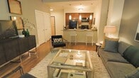 This single-floor residence is designed with an open-concept layout that connects the living area and kitchen, creating a cohesive and functional living environment. The living area features rich hardwood flooring, a plush area rug, and ample space for comfortable seating arrangements, anchored by a modern glass-top coffee table. Recessed and pendant lighting throughout the space provides a warm, inviting ambiance. The kitchen is a standout feature, showcasing warm-toned cabinetry, granite or stone countertops, and a peninsula with bar-height seating—ideal for casual dining or entertaining. A full suite of black appliances, including a refrigerator, microwave, and range, provides a sleek, contemporary contrast to the cabinetry. Pendant lights suspended above the peninsula add both style and task lighting. The open floor plan ensures seamless flow between the kitchen and living room, making the space feel expansive and connected. Double-door closets are visible near the entryway, offering generous storage. The neutral color palette, combined with warm wood tones and modern furnishings, gives the space a balanced, contemporary aesthetic that is both practical and visually appealing.