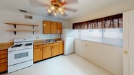 This single-floor unit offers a straightforward, functional layout connecting several well-defined living spaces. The kitchen is equipped with warm honey-toned wood cabinetry, open shelving with decorative iron brackets, a gas range, a double basin sink, and a white refrigerator. Vinyl flooring and a ceiling fan with a light fixture complete the kitchen, which also benefits from a window that allows natural light to fill the space.

The living area is generously proportioned, featuring neutral grey carpet, white walls, and direct sightlines into the kitchen and adjacent rooms. A six-panel closet door adds a traditional touch to the space.

The bedrooms are carpeted and bright, with white walls and windows dressed in sheer curtains. One bedroom includes a sliding closet with a hanging rod, offering practical storage. The rooms maintain a clean, open feel with ample wall space.

The bathroom features a shower-tub combination with tile surround, a pedestal-style sink, a mirrored medicine cabinet, and tile accents along the walls. The overall finishes are neutral and functional. Throughout the unit, the consistent use of grey carpet, white walls, and simple fixtures creates a cohesive, understated aesthetic.