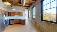 This thoughtfully converted industrial loft masterfully blends historic character with modern finishes across a single expansive floor. The open-plan living and kitchen area immediately impresses with its soaring timber beam ceiling, exposed silver ductwork, and a full wall of original yellow brick punctuated by large, arched black-framed windows that draw in abundant natural light and offer views of the surrounding cityscape. Light wood flooring runs throughout the main living area, reinforcing the warm, industrial aesthetic.

The well-appointed kitchen features warm wood cabinetry with sleek hardware, generous counter space, a double stainless steel sink, and a full suite of stainless steel appliances including a refrigerator, dishwasher, and range. Pendant lighting hangs from the timber ceiling, adding both function and character to the space.

The bedroom offers a more intimate setting, with plush carpeting, soft neutral walls, exposed beam accents above, and a spacious closet for ample storage. Two full bathrooms round out the unit — one featuring a tub/shower combo with a stacked washer and dryer, and the other showcasing a stone-accented accent wall, wood vanity, and large-format tile flooring. The overall ambiance is one of refined industrial charm, balancing raw architectural heritage with clean, contemporary living.