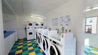 This single-floor laundry facility is designed for efficiency and ease of use. The room features a row of front-loading washing machines aligned along one wall, each equipped with individual water supply connections, providing ample capacity for multiple users at once. Complementing the washers is a bank of stacked dryer units, maximizing the use of vertical space and offering a generous number of drying options. The machines are maintained in excellent condition, presenting a clean, white aesthetic throughout. The walls are kept bright white, contributing to an open and hygienic atmosphere, and are accented by posted informational signage — including laundry tips and service instructions in both English and Spanish — ensuring accessibility for all users. Overhead fluorescent lighting provides clear, even illumination across the entire space. One of the most distinctive design elements is the bold, multicolored checkered floor, featuring a lively pattern of blue, green, and yellow tiles that adds a vibrant character to an otherwise utilitarian setting. The overall layout is practical and well-organized, making this laundry facility a straightforward and welcoming space for everyday use.