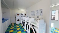 This single-floor laundry facility is designed for efficiency and ease of use. The room features a row of front-loading washing machines aligned along one wall, each equipped with individual water supply connections, providing ample capacity for multiple users at once. Complementing the washers is a bank of stacked dryer units, maximizing the use of vertical space and offering a generous number of drying options. The machines are maintained in excellent condition, presenting a clean, white aesthetic throughout. The walls are kept bright white, contributing to an open and hygienic atmosphere, and are accented by posted informational signage — including laundry tips and service instructions in both English and Spanish — ensuring accessibility for all users. Overhead fluorescent lighting provides clear, even illumination across the entire space. One of the most distinctive design elements is the bold, multicolored checkered floor, featuring a lively pattern of blue, green, and yellow tiles that adds a vibrant character to an otherwise utilitarian setting. The overall layout is practical and well-organized, making this laundry facility a straightforward and welcoming space for everyday use.