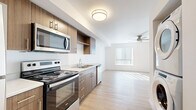Located at 10425 E Burnside St in Portland, OR, this efficiently designed single-floor unit offers a clean, contemporary aesthetic with thoughtful use of space. The open-concept layout seamlessly connects the kitchen and living area, creating a cohesive and functional living environment. The kitchen is well-appointed with warm wood-toned cabinetry, light quartz-style countertops, a double-basin sink, and a full suite of stainless steel appliances including an electric range, over-the-range microwave, refrigerator, and dishwasher. Upper cabinetry and open shelving provide practical storage solutions. The living area extends naturally from the kitchen, featuring light wood-style plank flooring that runs throughout the unit, crisp white walls, and a ceiling fan that enhances comfort and airflow. A large window dressed with vertical blinds allows ample natural light to pour in, brightening the space considerably. The bathroom continues the modern design language, showcasing a wood-toned vanity with a white countertop, a large mirror framed by overhead lighting, and the same light flooring. The overall ambiance is bright, clean, and move-in ready, reflecting a modern minimalist style suited for everyday living.