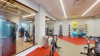 Located at 445 Hialeah Dr in Hialeah, FL, this contemporary fitness center is thoughtfully designed to accommodate a full range of workout activities across a single floor. The space is divided into distinct functional zones, each crafted to support a specific training focus.

The cardio area stands out with floor-to-ceiling windows that invite natural light and offer outdoor views of a lush poolside setting. This section houses a comprehensive lineup of modern equipment including treadmills, ellipticals, stationary bikes, and stair climbers, all set on warm wood-tone flooring. Exposed ductwork and linear pendant lighting lend the area an open, industrial-modern aesthetic.

Adjacent to the cardio zone, the strength training area features a robust selection of cable machines and resistance equipment arranged on matching hardwood-style flooring. Full-length mirrors amplify the sense of space and allow users to monitor their form. The exposed ceiling with visible HVAC infrastructure adds to the loft-like character of this section.

A dedicated cycling and functional training studio, separated by glass-panel partitions, features a vibrant red rubberized floor and is equipped with spin bikes, medicine balls, battle ropes, and weighted ball racks. The studio offers a dynamic, high-energy environment ideal for group or individual training sessions. Throughout the facility, bright linear lighting ensures excellent visibility in every zone.