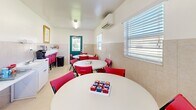Located at 1416 Griffin Rd in Leesburg, Florida, this single-room activity and game space offers a welcoming, functional environment designed for social interaction and leisure. The room is neatly organized around multiple round white tables surrounded by bright red folding chairs, creating a lively and inviting atmosphere throughout the space.

The interior features a classic two-tone wall treatment, with neutral tile wainscoting along the lower half of the walls transitioning to smooth white paint above, giving the room a clean and orderly appearance. Light tile flooring runs throughout, complementing the crisp, bright aesthetic. Natural light enters through multiple windows fitted with horizontal blinds, and a distinctive teal-painted entry door with a window adds a charming pop of color to the room.

A wall-mounted mini-split air conditioning unit ensures year-round comfort. Along one wall, a refreshment station includes a small white cabinet with a sink, a coffee maker, and stored beverages, making the space well-equipped for extended gatherings. A shelving unit and a counter with books and small decorative items add a personal touch. A framed botanical print in a gold frame adorns the wall, contributing a touch of warmth to the overall décor.

Game sets such as chess and tic-tac-toe are laid out on the tables, reinforcing this space's purpose as a dedicated area for recreational activities and community engagement.