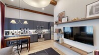 This well-appointed 1-bedroom, 1-bathroom residence makes exceptional use of its single-floor layout, combining modern design with carefully curated details across six distinct spaces. The living room is an immediate highlight, featuring soaring ceilings with exposed wood beams, light hardwood flooring, and tall windows dressed in rich burgundy drapes that flood the room with natural light. Built-in wood shelving provides both storage and display space, complementing the warm, eclectic aesthetic. The kitchen is sleek and fully equipped, showcasing floor-to-ceiling gray cabinetry with brushed copper hardware, white quartz countertops, a geometric tile backsplash, and a full suite of stainless steel appliances. The bedroom offers impressively tall ceilings and a calm, neutral palette, providing a restful retreat. The bathroom is refined and spa-like, featuring large-format tile flooring, a floating wood vanity with an integrated white basin, matte black fixtures, and a glass-enclosed walk-in shower with dramatic floor-to-ceiling tile surround. A hallway connects the spaces and is accented with vintage-style artwork, adding character. A utility room rounds out the functional layout of this efficient yet stylish residence.