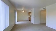 This single-floor space offers an open and functional layout that seamlessly connects the main living area to the kitchen. The living area is finished with wall-to-wall carpet in a neutral beige tone, creating a warm and understated foundation throughout the space. A ceiling fan with a light fixture is centrally positioned overhead, providing both air circulation and ambient lighting to the room.

The walls are painted in a soft, light neutral tone that enhances the brightness of the interior. Visible air vents integrated into the walls and ceiling indicate a central HVAC system for year-round climate control.

The kitchen is partially open to the living area and features wood-toned cabinetry, white appliances including a microwave and range, and light countertops. The layout appears compact yet efficient, with cabinetry providing ample storage. A doorway adjacent to the kitchen leads to an additional room or hallway, and a second door is visible on the right side of the living area, suggesting access to further rooms within the unit. The overall ambiance is clean, simple, and move-in ready.