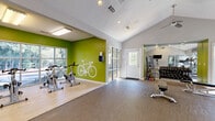 Located at 11251 Alpharetta Hwy in Roswell, GA, this fitness center offers a comprehensive and energizing workout environment spread across a single floor. The facility is divided into distinct functional zones, each designed to support a variety of exercise routines. The main cardio area features multiple treadmills and elliptical machines positioned along a wall of large windows, flooding the space with natural light and offering outdoor views of the surrounding greenery. A bold "MAKE IT HAPPEN." wall decal above the windows sets an motivating tone throughout. The free weights area is well-equipped with a full dumbbell rack, kettlebells, and an adjustable weight bench, complemented by floor-to-ceiling mirrors that enhance the sense of space. A dedicated group exercise and functional training studio showcases soaring vaulted ceilings with a ceiling fan, carpeted flooring, stability balls, TRX suspension straps, and a wall-mounted TV — all anchored by an eye-catching "WAKE. RUN. LIFT. EAT. SLEEP. REPEAT." motivational graphic. The spin cycling zone features a vibrant lime-green accent wall adorned with a white bicycle silhouette, housing multiple stationary cycling bikes on light hardwood-style flooring. Natural light permeates throughout, creating a bright, welcoming, and high-energy atmosphere.