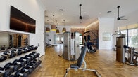 This fitness center offers a thoughtfully designed, multi-zone workout environment that balances form and function. The space is unified by warm parquet hardwood flooring throughout and crisp white walls adorned with crown molding, creating a polished yet welcoming ambiance. Stylish pendant lights with oversized drum shades complement recessed ceiling lighting, while ceiling fans contribute to a comfortable atmosphere during workouts.

A standout feature is the exposed brick accent wall, which adds a distinctive, textured contrast to the otherwise clean, modern interior. The cardio zone is bathed in natural light from floor-to-ceiling windows fitted with roller shades, offering views of lush outdoor greenery. This area houses a full row of True-brand treadmills, ellipticals, and stationary bikes for comprehensive aerobic training.

The strength training area is equally well-equipped, featuring Cybex cable machines, a multi-functional cable rack, weight benches, and a full dumbbell rack. A separate room accessible through glass-paned French doors accommodates indoor cycling bikes, providing a dedicated space for spin workouts. Wall-mounted flat-screen televisions are positioned throughout for entertainment. The facility's generous proportions, abundant equipment, and refined architectural details combine to deliver a premium fitness experience.