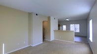 This single-floor space is designed with an open-concept layout that promotes a sense of spaciousness and easy flow between living areas. The main living area features neutral-toned carpet underfoot and warm beige walls, creating a clean, understated aesthetic throughout.

A defining architectural feature is the half-wall peninsula that separates the living space from the kitchen area, providing visual distinction between the two zones while maintaining an open and connected feel. The kitchen sits adjacent to the living area and benefits from abundant natural light streaming through large windows, brightening the space considerably.

Ceiling-mounted lighting fixtures provide additional illumination in the kitchen area, while HVAC vents integrated into the walls and ceiling ensure climate control throughout. A hallway accessible from the main living area leads to additional rooms, offering privacy from the common spaces.

White baseboards provide a crisp contrast against the beige walls and carpeted flooring, contributing to the overall clean and well-maintained appearance of the interior. The single-floor layout ensures accessibility and practicality, making the space straightforward to navigate.
