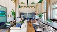 Located at 3015 N Rocky Point Dr E in Tampa, Florida, this single-floor amenity space delivers an impressive blend of modern design and upscale functionality. The layout is anchored by dramatic double-height ceilings that amplify the sense of openness, while floor-to-ceiling windows and glass doors flood the interior with natural light and offer views of the surrounding outdoor scenery.

The kitchen is a standout feature, outfitted with custom gray shaker cabinetry accented by brushed gold hardware, a striking textured green tile backsplash, stainless steel appliances including a French door refrigerator, and a gas range. Warm wood-look tile flooring and globe pendant lights in a brushed gold finish add to the refined atmosphere.

The open lounge area showcases a grand white marble-veined island with seating, complemented by a mix of high-top tables with marble tops and tufted leather seating on brushed gold bases. Ornate drum chandeliers hang from the soaring teal-accented ceiling, while lush tropical plants bring life to every corner. Large windows and glass entry doors create a seamless connection to the outdoors, making this a bright, welcoming, and beautifully appointed communal space.