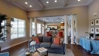 This single-story space showcases an open-concept design that integrates the living, dining, and kitchen areas into one cohesive, flowing environment. The living area is anchored by a plush gray sectional sofa accented with warm coral and patterned throw pillows, complementing the rich hardwood flooring beneath. A large area rug defines the seating zone, which centers around a stone-topped coffee table styled with decorative accessories.

Distinctive white columns frame the transition between the living and dining spaces, adding an architectural elegance that draws the eye upward to the coffered ceiling detail overhead. Recessed lighting throughout provides consistent, well-distributed illumination, while multiple windows along the walls invite ample natural light into the space.

The dining area features coral-upholstered chairs around a central table, echoing the warm accent tones found throughout. Beyond, the kitchen is outfitted with white cabinetry, stainless steel appliances including a refrigerator and microwave, and a bold orange accent wall that energizes the space. A ceiling fan is visible in the kitchen zone, adding both function and a casual touch. The overall ambiance is warm, inviting, and thoughtfully coordinated.