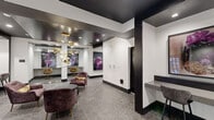 Located at 401 E Oglethorpe Ave in the heart of Savannah, Georgia, this single-floor residential lobby serves as a sophisticated and welcoming entrance to the building. The space is thoughtfully designed with a striking contrast of crisp white walls and bold dark ceilings, creating a contemporary aesthetic that immediately sets an upscale tone.

The lounge area is anchored by plush, deep-mauve velvet armchairs arranged around a sleek dark coffee table, providing a comfortable and stylish gathering spot for residents. A sculptural gold chandelier with cascading globe lights serves as a dramatic focal point overhead, complementing the warm gold accents found throughout the space. Large-scale abstract artwork in deep purples and magentas adorns the walls, adding a bold artistic character to the interior.

A dedicated mail center features an extensive array of individual bronze mailboxes mounted both on a central island unit and along the wall, efficiently serving the building's residents. Glass-partitioned alcoves along one side of the lobby offer semi-private workstation nooks with counter seating, ideal for quick tasks or remote work. A built-in counter with bar-height seating near the entrance provides additional workspace.

Patterned grey tile flooring runs throughout, tying the various functional zones together cohesively. Large glass entry doors allow natural light to filter into the lobby, further brightening the refined and well-appointed space.
