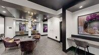 Located at 401 E Oglethorpe Ave in the heart of Savannah, Georgia, this single-floor residential lobby serves as a sophisticated and welcoming entrance to the building. The space is thoughtfully designed with a striking contrast of crisp white walls and bold dark ceilings, creating a contemporary aesthetic that immediately sets an upscale tone.

The lounge area is anchored by plush, deep-mauve velvet armchairs arranged around a sleek dark coffee table, providing a comfortable and stylish gathering spot for residents. A sculptural gold chandelier with cascading globe lights serves as a dramatic focal point overhead, complementing the warm gold accents found throughout the space. Large-scale abstract artwork in deep purples and magentas adorns the walls, adding a bold artistic character to the interior.

A dedicated mail center features an extensive array of individual bronze mailboxes mounted both on a central island unit and along the wall, efficiently serving the building's residents. Glass-partitioned alcoves along one side of the lobby offer semi-private workstation nooks with counter seating, ideal for quick tasks or remote work. A built-in counter with bar-height seating near the entrance provides additional workspace.

Patterned grey tile flooring runs throughout, tying the various functional zones together cohesively. Large glass entry doors allow natural light to filter into the lobby, further brightening the refined and well-appointed space.