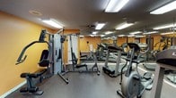 This single-floor fitness center is designed to provide a comprehensive workout environment in a compact, functional layout. The space is defined by its bold amber-orange walls, textured ceilings, and flush fluorescent panel lighting that ensures consistent, even illumination throughout. Full-wall mirrors line multiple sides of the main workout area, visually expanding the space and allowing users to monitor their form during exercise. The flooring features durable, rubber-style material well-suited for a high-traffic gym environment. The cardio section is equipped with Landice treadmills and an elliptical cross-trainer, offering a range of aerobic workout options. A multi-station cable strength machine anchors the free weights area, accompanied by an adjustable bench for versatile resistance training. A water cooler station is conveniently positioned along one wall, providing easy hydration access. Entry to the space is through a black-framed glass door, with a fire extinguisher mounted nearby for safety compliance. The overall layout is efficient and accessible, making it a practical facility for a variety of fitness routines.