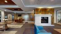 Located at 100 Exchange St in Malden, MA, this single-floor amenity space is thoughtfully designed to offer residents a well-rounded mix of comfort, productivity, and wellness. The welcoming lobby sets an elegant tone with rich wood-paneled walls, a wall-mounted television, a reception desk, and a striking marble-surround fireplace flanked by contemporary sconce lighting. Vibrant blue and orange seating arrangements create a warm, inviting atmosphere throughout the common lounge areas, which are accented by patterned carpeting and recessed tray ceilings. The co-working area provides a bright, open environment for productivity, featuring multiple white collaborative tables, ergonomic seating in a mix of colors, and generous natural light from large windows. The space also includes a kitchenette with counter seating and stainless steel appliances, as well as colorful artwork displayed throughout. The fitness center is expansive and well-equipped, offering cardio machines including treadmills, ellipticals, and stair climbers, all positioned beneath a tray ceiling with views through large windows. A dedicated strength training area features a multi-use cable rack, free weights, battle ropes, and medicine balls. Glass partitions visually connect the fitness areas to the lounge, enhancing the open, integrated feel of the floor.