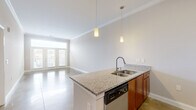 Located at 404 W Smith St in Greensboro, NC, this one-bedroom, one-bathroom residence offers a well-designed single-floor layout that maximizes functionality and style. The open-concept kitchen and living area serves as the heart of the home, featuring a central island with granite countertops, a double-basin sink, and pendant lighting overhead. The kitchen is equipped with warm wood cabinetry, a subway tile backsplash, and a full suite of stainless steel appliances including a refrigerator, gas range, microwave, and dishwasher. Polished concrete floors extend throughout the main living area, adding an industrial-modern aesthetic, while crown molding lends a refined architectural detail. The living room is flooded with natural light through French doors topped with a transom window, offering outdoor views and a bright, airy ambiance. The bedroom is carpeted for comfort, includes a ceiling fan, and features a window with views to the outside. Two closets provide ample storage, including a spacious walk-in with wire shelving. The bathroom is finished with tile flooring and includes a tub/shower combination. A dedicated hallway connects the private and shared spaces efficiently, completing this thoughtfully laid-out residence.