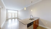 Located at 404 W Smith St in Greensboro, NC, this one-bedroom, one-bathroom residence offers a well-designed single-floor layout that maximizes functionality and style. The open-concept kitchen and living area serves as the heart of the home, featuring a central island with granite countertops, a double-basin sink, and pendant lighting overhead. The kitchen is equipped with warm wood cabinetry, a subway tile backsplash, and a full suite of stainless steel appliances including a refrigerator, gas range, microwave, and dishwasher. Polished concrete floors extend throughout the main living area, adding an industrial-modern aesthetic, while crown molding lends a refined architectural detail. The living room is flooded with natural light through French doors topped with a transom window, offering outdoor views and a bright, airy ambiance. The bedroom is carpeted for comfort, includes a ceiling fan, and features a window with views to the outside. Two closets provide ample storage, including a spacious walk-in with wire shelving. The bathroom is finished with tile flooring and includes a tub/shower combination. A dedicated hallway connects the private and shared spaces efficiently, completing this thoughtfully laid-out residence.