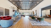 This expansive, single-floor amenity space is designed to impress, blending contemporary style with warm, inviting details throughout. The main lounge area commands attention with soaring ceilings, a dramatic floor-to-ceiling stone accent wall housing a linear fireplace, and elegant chandelier lighting. Wood-look flooring runs throughout, anchored by bold patterned area rugs that define distinct seating zones furnished with plush sofas, velvet armchairs, and curated artwork. A grid-panel ceiling with recessed lighting adds an industrial-modern contrast to the bright white walls.

The kitchen and bar area features a long white quartz island with upholstered counter seating, marble-look tile backsplash, dark cabinetry, and striking geometric gold pendant lights — ideal for social gatherings and community events. Adjacent dining and collaborative seating areas offer flexible configurations with designer chairs and patterned rugs beneath high ceilings with French door access to the outdoors.

The dedicated game room stands out with richly textured exposed wood beam ceilings, a full-size billiards table, shuffleboard table, and additional lounge seating, all set against the same stone wall feature. Private office space with glass sliding doors and built-in shelving rounds out this well-appointed, multifunctional community facility.