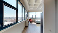 This single-floor unit is situated on an upper level, offering sweeping panoramic views of the surrounding cityscape through expansive teal-framed windows that line multiple walls. The architectural design features distinctive coffered ceilings throughout, lending a structured, industrial-modern aesthetic that sets the space apart.

The open-concept living and kitchen area flows seamlessly, with light wood-tone flooring underfoot and generous natural light streaming in from large windows. The kitchen is well-appointed with white cabinetry extending to the ceiling, stainless steel appliances including a full-size refrigerator and built-in microwave, a single-basin sink, and a butcher block-topped island cart that provides additional prep and storage space.

The bedroom is a standout feature, wrapped on two sides by floor-to-ceiling windows that frame expansive views of the city skyline and infrastructure below. The coffered ceiling continues here, enhancing the sense of openness and architectural character.

The bathroom is clean and functional, finished with wood-tone flooring, a white vanity with under-cabinet storage, a mirror with overhead lighting, and a full bathtub with shower. The overall ambiance of the unit is bright, airy, and contemporary.