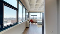 This single-floor unit is situated on an upper level, offering sweeping panoramic views of the surrounding cityscape through expansive teal-framed windows that line multiple walls. The architectural design features distinctive coffered ceilings throughout, lending a structured, industrial-modern aesthetic that sets the space apart.

The open-concept living and kitchen area flows seamlessly, with light wood-tone flooring underfoot and generous natural light streaming in from large windows. The kitchen is well-appointed with white cabinetry extending to the ceiling, stainless steel appliances including a full-size refrigerator and built-in microwave, a single-basin sink, and a butcher block-topped island cart that provides additional prep and storage space.

The bedroom is a standout feature, wrapped on two sides by floor-to-ceiling windows that frame expansive views of the city skyline and infrastructure below. The coffered ceiling continues here, enhancing the sense of openness and architectural character.

The bathroom is clean and functional, finished with wood-tone flooring, a white vanity with under-cabinet storage, a mirror with overhead lighting, and a full bathtub with shower. The overall ambiance of the unit is bright, airy, and contemporary.