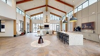 This expansive single-floor communal space makes an immediate impression with its dramatic vaulted ceilings supported by exposed natural wood beams, creating a grand, lodge-inspired atmosphere. Towering floor-to-ceiling windows and clerestory glazing wrap the space, flooding the interior with abundant natural light and offering open views of the surrounding exterior. The large-format stone tile flooring anchors the space with an earthy, neutral palette that complements the warm wood tones throughout.

At the entrance, a striking marble-clad reception desk serves as a commanding focal point, accented by a stylish multi-arm chandelier overhead. The entry lounge features a classic tufted leather Chesterfield sofa, a patterned area rug, and suspended wooden bench seating positioned beside the expansive glazed entry doors.

The central open area is defined by a sculptural pedestal accent table set on a round rug, drawing the eye toward a comfortable seating arrangement anchored by a fireplace and wall-mounted television. An antler chandelier adds a rustic, organic touch above. Adjacent to this lounge area is a long quartz-topped bar counter with black barsttools, a coffee station, and cabinetry, providing a functional social hub. The overall ambiance blends contemporary sophistication with warm, natural materials for an inviting and well-appointed communal environment.