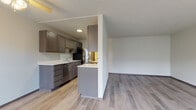 This single-floor unit offers a practical, open-concept layout with consistent light wood-look flooring running throughout. The kitchen is well-appointed with dark taupe cabinetry offering generous storage, white countertops, stainless steel appliances including a dishwasher and refrigerator, and a peninsula that separates the cooking space from the adjoining living area. A ceiling fan with a light fixture overhead serves both the kitchen and the open living space, which is defined by a partial dividing wall. The living area is bright and open, featuring a large picture window that allows natural light to fill the room and provides a view of the surrounding exterior. The textured ceiling is consistent throughout the main areas. The bathroom features matching dark taupe cabinetry, a white undermount sink with a modern faucet, a large mirror, and clean white walls, maintaining the cohesive design palette found throughout the unit. A hallway connects the bathroom to the rest of the space, with glimpses of additional storage and in-unit laundry connections visible. Overall, the unit presents a clean, updated aesthetic with a neutral color palette suited to a variety of living arrangements.