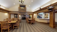 This single-story community is thoughtfully designed to support an active and comfortable lifestyle. The centerpiece is a grand common dining area distinguished by soaring vaulted ceilings, decorative wood trim with an ornamental border, and a striking interior balcony with a chandelier overhead. Wood-crafted columns frame the space, which accommodates multiple dining tables and chairs arranged for communal gatherings. An adjacent open kitchen pass-through allows for efficient meal service. A upright piano and lounge seating are visible in the background, adding a social and recreational dimension to the common area. A dedicated information nook features a bulletin board, brochure display shelving, and resident mailboxes, serving as a practical communication hub. The on-site salon is well-appointed with a professional styling chair, large mirror, product shelving, and a cozy seating area. A computer room offers multiple workstations along a built-in wraparound desk, ideal for residents staying connected. The fitness room is equipped with treadmills and cardio equipment, accented by bold wall colors and natural light from a window. Throughout, warm tones, decorative borders, and wood accents create a cohesive and inviting atmosphere.