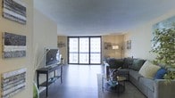 This single-floor residence offers a clean, modern living environment centered around a well-appointed open-plan living area. The space is anchored by large sliding glass doors that flood the interior with natural light and provide direct access to a balcony, framing views of the surrounding urban landscape. The smooth, light-toned walls complement the dark flooring, creating a stylish contrast that gives the space a contemporary feel.

The living area is thoughtfully arranged, featuring a sectional sofa positioned around a pair of round glass coffee tables atop a textured area rug, creating a comfortable and inviting conversation area. A sleek media console with a flatscreen television occupies one side of the room, while an arc floor lamp adds warm ambient lighting near the seating area. Decorative wall art and accent pieces add personality throughout the space without overwhelming the clean aesthetic.

The textured ceiling adds subtle architectural character, and the open layout allows for a natural flow between functional zones. Overall, the space presents a balanced combination of modern design and practical comfort, making it a well-lit, stylish, and functional living environment suited for everyday living and entertaining alike.