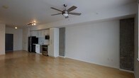 This single-floor space showcases a thoughtfully designed open-concept layout that seamlessly integrates the living area and kitchen into one cohesive environment. Light hardwood flooring extends throughout, lending warmth and continuity to the space while reflecting available light to create a bright, airy atmosphere.

A prominent ceiling fan with brushed nickel finishing is centrally mounted, providing both functional air circulation and a subtle modern design accent. The clean white walls offer a neutral backdrop, making the space feel expansive and versatile.

The kitchen is positioned along one wall and features stainless steel appliances, including a refrigerator, range, and microwave. White cabinetry with open shelving above provides practical storage while maintaining a contemporary aesthetic. A stainless steel sink sits beneath task lighting, and a distinct concrete or stone accent column serves as a visual divider between the kitchen and the open living area, adding an industrial design element that contrasts effectively with the otherwise clean, minimalist interior.

Track lighting near the kitchen enhances task functionality, while the overall layout maximizes usable floor space throughout.