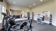 This fitness center offers a well-rounded workout environment designed to accommodate a variety of exercise routines. The single-level space is outfitted with durable carpet flooring and illuminated by recessed ceiling lighting, creating a bright and functional atmosphere conducive to physical activity.

The cardio section features multiple treadmills and a recumbent exercise bike, providing options for low- and high-impact cardiovascular training. Alongside these, a cable machine and a dedicated strength machine offer versatile resistance training capabilities, while a rack of dumbbells lines the mirrored wall for free-weight exercises. A weight bench is also available for additional strength training.

A large floor-to-ceiling mirror spans one wall, enhancing the sense of space and allowing users to monitor their form during workouts. A wall-mounted air conditioning unit ensures comfortable temperatures throughout the room. The neutral, light-toned walls contribute to the clean and open feel of the space. An adjacent doorway suggests access to additional rooms or facilities, adding to the overall convenience of this well-appointed fitness center.
