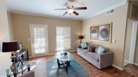 This single-floor residence offers a thoughtfully designed layout that balances comfort and functionality. The living room serves as the heart of the home, featuring warm hardwood-style flooring, crown molding, and a ceiling fan with light fixture. Multiple windows and a glass-paned exterior door flood the space with natural light, while neutral beige walls create a welcoming ambiance throughout.

The kitchen is equipped with dark cabinetry, black appliances, and a peninsula-style breakfast bar with a durable countertop, offering a practical workspace and casual dining option. A louvered closet door provides additional storage access nearby.

Two well-sized bedrooms offer comfortable retreats, each with plush carpeting and ceiling fans. One bedroom features a distinctive arched window that frames outdoor greenery, while the other benefits from abundant natural light through multiple windows. Both rooms include closet space, with at least one walk-in closet visible.

The two bathrooms are cleanly appointed — one features a tiled walk-in shower with glass sliding doors, while the other includes a shower-tub combination with a striped curtain, a single vanity with a broad mirror, and tasteful wall art. The consistent warm palette and thoughtful details throughout the space create a cohesive and inviting living environment.