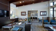 This single-story communal space exemplifies contemporary design with a thoughtful blend of warm and modern elements. The interior is immediately distinguished by its striking wood-paneled ceiling with exposed beams and recessed lighting, contrasting elegantly against crisp white walls to create a refined, layered aesthetic.

The main lounge area is generously proportioned, featuring a carefully curated seating arrangement of plush gray sofas, lounge chairs, and low-profile coffee tables arranged atop patterned carpeting. Woven rattan privacy screens add texture and subtle definition to seating zones, while framed artwork and decorative floral arrangements contribute visual interest and warmth throughout.

Large, multi-pane black-framed windows line the perimeter, flooding the interior with abundant natural light and offering views of the surrounding landscape. Near the entrance, glass double doors enhance the welcoming atmosphere.

An adjoining dining or conference room, visible through an interior opening, features a full dining table with dark chairs and a distinctive round sculptural wall installation, providing a functional yet stylish secondary gathering space. A credenza with cabinetry anchors one wall, adding practical storage while complementing the overall design scheme. The space projects a polished, hospitality-inspired ambiance throughout.