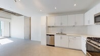 This single-floor space showcases a contemporary, minimalist design with an open-concept layout that maximizes the sense of space and flow. The kitchen is a standout feature, outfitted with flat-front white cabinetry, light stone countertops, a white subway tile backsplash, and a full suite of stainless steel appliances including a gas range, over-the-range microwave, dishwasher, and refrigerator. Recessed lighting illuminates the kitchen and living areas efficiently throughout.

The living area benefits from soaring ceilings with exposed raw concrete soffits that lend an industrial-modern character to the space. Large sliding windows allow generous natural light to fill the room, complemented by neutral concrete-finish flooring that runs throughout. An accent wall of exposed concrete adds texture and an urban edge to the otherwise clean, white interior.

The bathroom is thoughtfully appointed with a white vanity, integrated sink, large mirror with overhead lighting, and a glass-enclosed shower featuring elegant tile surround in warm tones. The overall ambiance of the space is clean, bright, and contemporary — ideal for those who appreciate modern architecture with industrial design influences.
