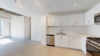 This single-floor space showcases a contemporary, minimalist design with an open-concept layout that maximizes the sense of space and flow. The kitchen is a standout feature, outfitted with flat-front white cabinetry, light stone countertops, a white subway tile backsplash, and a full suite of stainless steel appliances including a gas range, over-the-range microwave, dishwasher, and refrigerator. Recessed lighting illuminates the kitchen and living areas efficiently throughout.

The living area benefits from soaring ceilings with exposed raw concrete soffits that lend an industrial-modern character to the space. Large sliding windows allow generous natural light to fill the room, complemented by neutral concrete-finish flooring that runs throughout. An accent wall of exposed concrete adds texture and an urban edge to the otherwise clean, white interior.

The bathroom is thoughtfully appointed with a white vanity, integrated sink, large mirror with overhead lighting, and a glass-enclosed shower featuring elegant tile surround in warm tones. The overall ambiance of the space is clean, bright, and contemporary — ideal for those who appreciate modern architecture with industrial design influences.