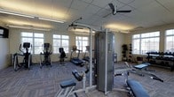 This fitness center is a thoughtfully appointed exercise facility designed to support a wide range of workout routines. The space is bathed in natural light streaming through multiple large windows along the walls, creating an open and energizing atmosphere. A drop ceiling with recessed lighting and a ceiling fan ensure the environment remains well-lit and comfortable during any workout session.

The cardio area is equipped with a treadmill, an elliptical trainer, and a stationary bike, offering users a variety of options for cardiovascular exercise. A prominent multi-station cable machine serves as a centerpiece for strength training, providing versatility for targeting multiple muscle groups. Additional strength equipment includes a dedicated bench and a rack of medicine balls and weighted balls, catering to both traditional and functional fitness training.

The flooring features durable, patterned carpet tiles well-suited for a gym environment, offering cushioning and stability underfoot. Decorative potted plants add a touch of greenery and warmth to the space. The overall layout is organized and spacious, allowing for comfortable movement between equipment. This facility provides a comprehensive and accessible fitness environment suitable for a broad range of fitness levels and training goals.