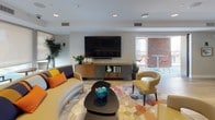 This well-appointed clubroom is a single-floor communal space designed to foster social interaction and relaxed gatherings. The layout is open and expansive, anchored by wide-plank light oak hardwood flooring that runs throughout and contributes to a warm, contemporary aesthetic.

The main lounge area features a curved sectional sofa and coordinating accent chairs arranged around a sculptural coffee table, all set atop a vibrant geometric-patterned area rug. Pops of teal, orange, and yellow in the furnishings add energy and personality to the otherwise neutral palette of soft grays and whites. Recessed lighting provides even illumination across the space.

A long, white-topped kitchenette counter with dark cabinetry serves as both a functional prep area and an informal bar, complete with bar-height seating. Built-in appliances are integrated below the counter, and a large wall-mounted flat-screen television with a soundbar anchors the media area nearby. Framed artwork and decorative objects add a curated, residential feel.

A secondary seating nook with a teal sofa and small café table offers a more intimate setting, while glass double doors lead to a private meeting or overflow room. Sliding glass doors open to an outdoor terrace with high-top bistro seating, extending the usable space and bringing in natural light.