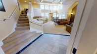 Located in Lakewood, Washington, this leasing office offers a polished and welcoming environment designed to serve residents and prospective tenants alike. The entry foyer makes an immediate impression with its soaring ceilings, arched transom windows, elegant chandelier, and light tile flooring accented with navy blue inlays — setting a refined tone from the moment visitors arrive.

Framed glass French doors lead from the foyer into the main common area, where the open layout connects a comfortable lounge, dining space, and kitchenette. The lounge is anchored by a striking full brick fireplace framed by an octagonal arch, flanked by floor lamps, and surrounded by plush seating arranged around wooden coffee tables. Natural light pours in through an abundance of large windows, particularly in the sunlit dining area, which features high clerestory windows on multiple sides and a casual dining set beneath a chandelier.

The kitchenette includes a breakfast bar with bar seating and dark cabinetry, providing a convenient space for staff and guests. A staircase with white railings and an upper landing adds architectural depth to the interior. Throughout the space, warm yellow tones, carpeted flooring in the lounge areas, and tile in transitional zones create a cohesive and inviting atmosphere suited for a professional leasing environment.