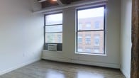 This single-floor space showcases a clean, open layout with notable urban character. Large double-hung windows dominate the exterior wall, flooding the interior with natural light while offering views of the surrounding brick architecture — a hallmark of classic city streetscapes. The windows are equipped with window-unit air conditioning, providing climate control alongside baseboard heating that runs the length of the wall for year-round comfort. The flooring consists of hardwood planks in a warm, medium tone, lending a polished yet approachable feel to the space. Overhead, the ceiling reveals exposed industrial elements, including visible beams and structural components that add architectural interest and a loft-like aesthetic. The walls are finished in a neutral off-white, creating a versatile backdrop suitable for a variety of uses. The overall ambiance balances urban industrial character with functional simplicity, making the space adaptable for residential or light commercial purposes. The generous window presence ensures the interior feels bright and connected to the city environment outside.