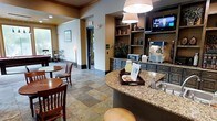 This single-floor hospitality space combines a functional service area with a relaxed lounge, designed to accommodate guests comfortably. At its centerpiece is a curved granite-topped counter with an integrated sink, providing a practical refreshment station equipped with beverage dispensers and snack service. Pendant lighting hangs above the counter, casting a warm glow over the polished stone surface.

Behind the counter, dark-toned cabinetry with granite countertops houses small appliances, including a toaster oven and popcorn machine, while open built-in shelving above displays decorative accents and a wall-mounted flat-screen television. The shelving unit adds both visual depth and functionality to the service zone.

The lounge area features slate tile flooring in earthy, multi-toned hues, lending a grounded, natural feel to the space. Wooden dining chairs with upholstered seats surround round tables, creating an inviting setting for guests to gather. A floor lamp in the corner adds a residential warmth to the atmosphere.

Natural light filters in through a glass-paneled door, while additional wall art and neutral paint tones in warm beige and amber contribute to the space's welcoming, unpretentious character.